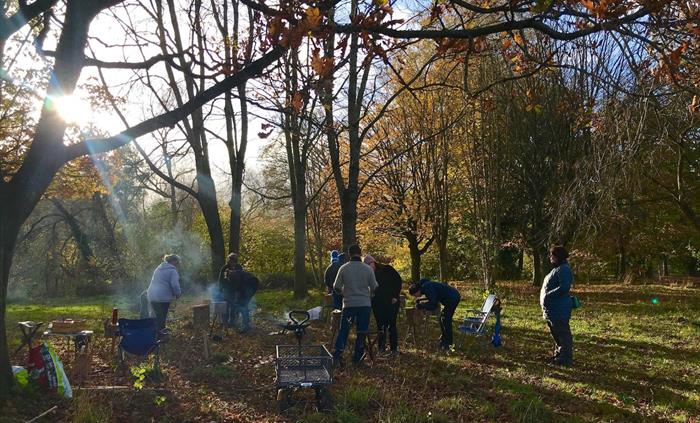 People standing in a circle in a small copse each with their own  2ft upright log as a workstation for carving. Surrounded on left and behind by trees